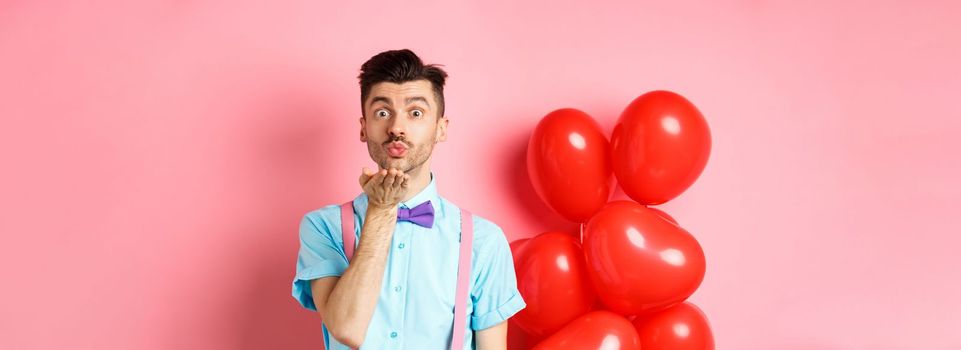 Valentines day concept. Attractive and funny man in bow-tie, blowing air kiss to lover, standing near pink background and red hearts balloons.