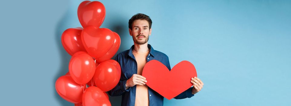 Valentines day and love concept. Young man showing red paper heart cutout and standing near romantic balloons, looking at lover, standing over blue background.
