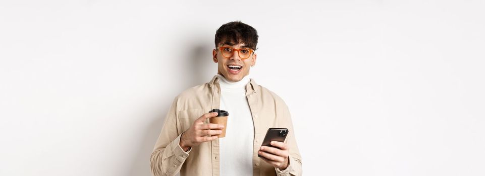 Cheerful smiling young man in glasses drinking coffee and holding smartphone, looking excited at camera, standing on white background.