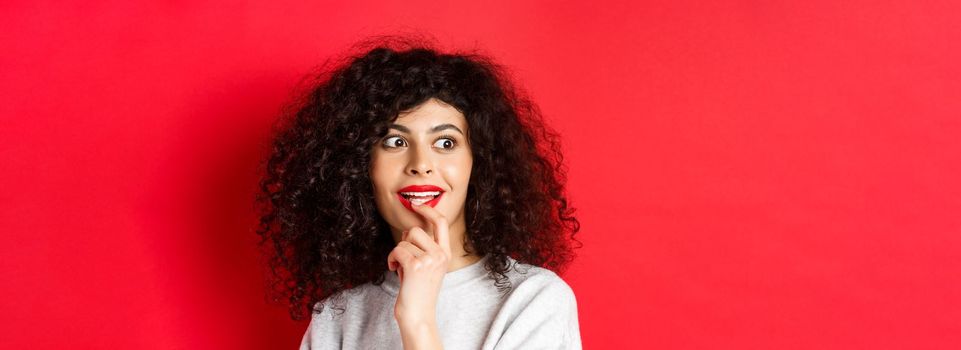 Close-up of excited curly woman touching lips and looking aside with interest, checking out cool promo, standing against red background.