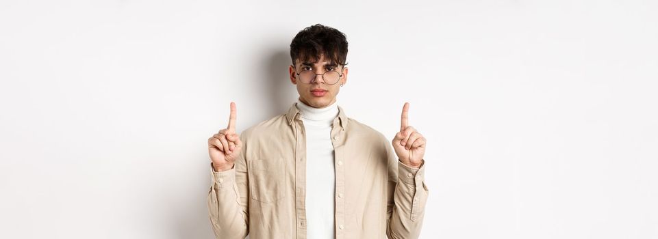 Handsome hipster guy in glasses pointing fingers up at empty space, looking serious at camera, standing on white background.