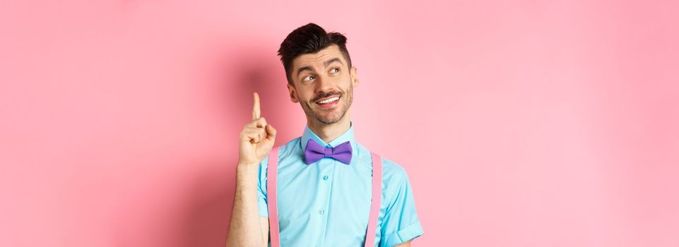 Handsome smiling man raising finger in eureka gesture, looking away and pitching an idea, have suggestion or plan, standing in bow-tie over pink background.