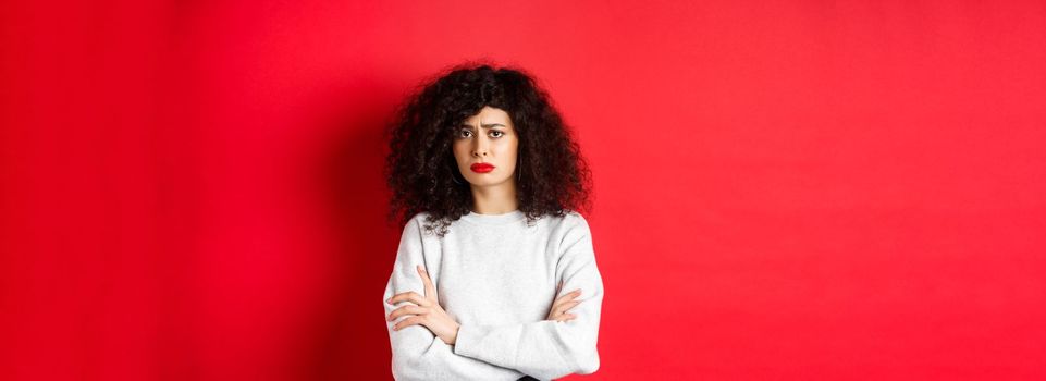 Sad and worried caucasian woman frowning, cross arms on chest and looking concerned, feeling bad, standing on red background.