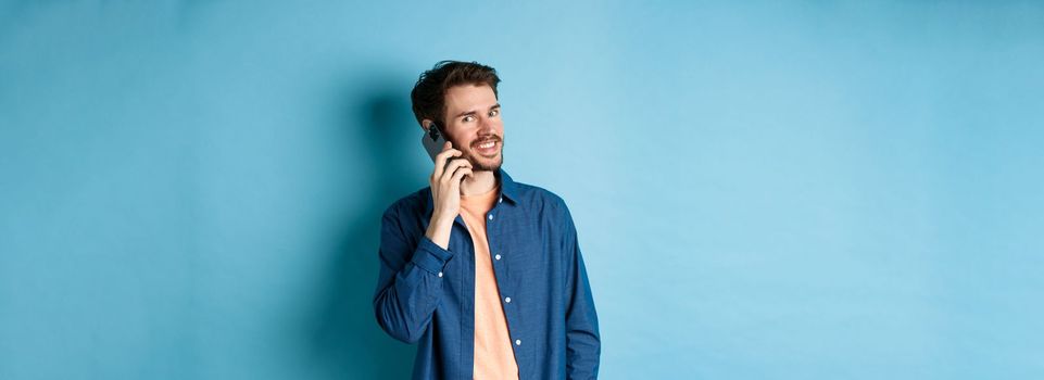 Modern smiling guy talking on mobile phone, looking happy at camera, standing on blue background.