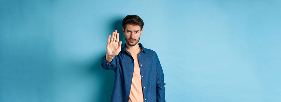 Upset man frowning and asking to stop, stretch out hand to prohibit or disagree with something bad, standing on blue background.