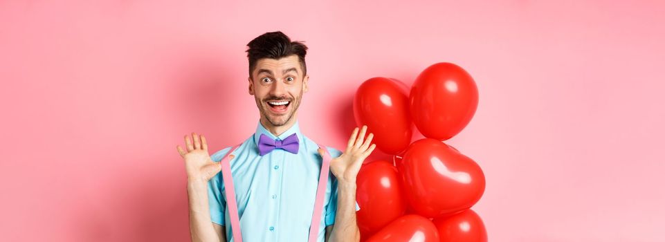 Valentines day concept. Happy young man looking surprised, raising hands up joyful, celebrating near big red hearts and pink background.