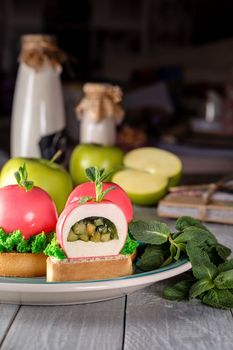 Light mousse dessert in shape of apples covered with mirror pink glaze, stuffed with natural fruit compote on shortbread tartlets with caramel, walnuts layer, green biscuit grass and fresh mint leaves