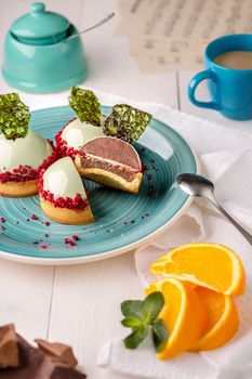 Delicious sugar dough tartlets with sourish orange confit, layer of nut crumbs and hemisphere of sweet airy chocolate mousse covered with glossy vanilla icing decorated with colored caramel on plate