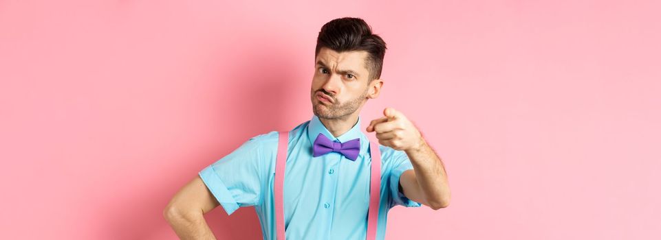 Grumpy guy in bow-tie and suspenders accusing you, its your fault gesture, pointing finger at camera with disappointed frowning face, pink background.