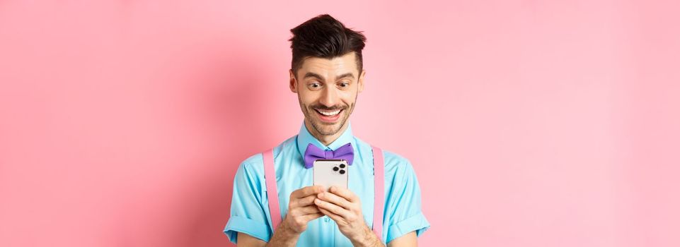 Online shopping. Happy guy looking at smartphone screen, reading message and smiling, standing on pink background.