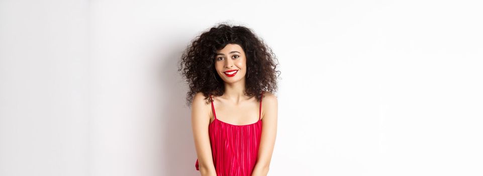 Cute modest lady in red dress and makeup, smiling and blushing, looking at camera, standing over white background.