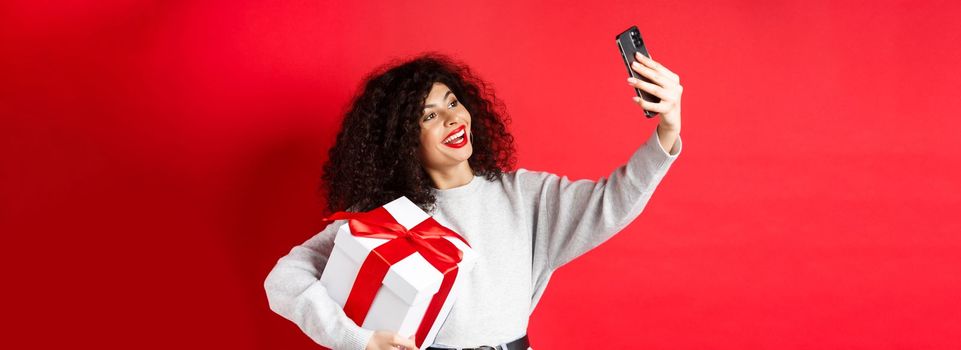 Holidays and tech concept. Happy woman taking selfie with her gift, holding present and smartphone, standing on red background.