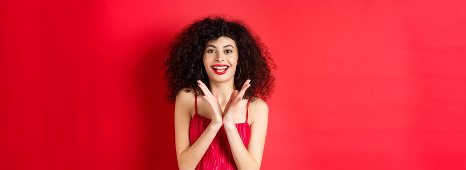 Cheerful young woman with curly hair, wearing evening dress, clap hands and smiling, praise good performance with applause, standing against red background.