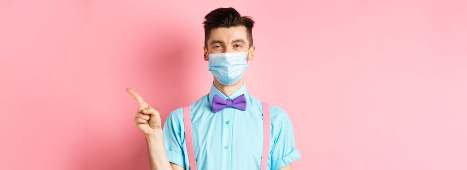 Covid-19, pandemic and health concept. Smiling caucasian man in medical mask pointing finger right, showing advertisement, standing on pink background.