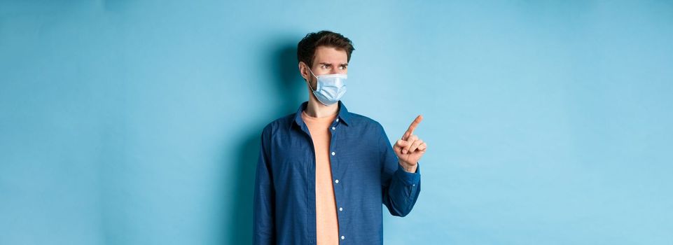 Covid-19 and healthcare concept. Confused and doubtful man in face mask frowning, pointing and looking at empty space, standing on blue background.