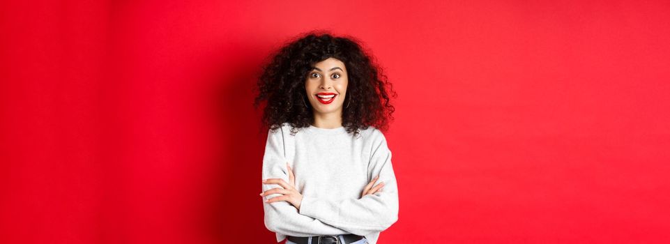 Cheerful young woman with curly hairstyle, raising eyebrows and looking surprised, hear interesting news, red background.