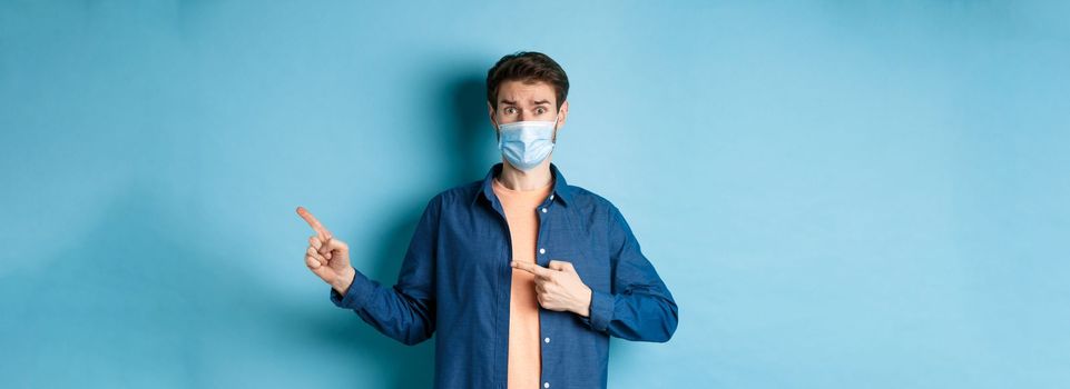 Covid and healthcare concept. Worried young man in medical mask raising eyebrow, pointing finger right with doubtful and nervous face, standing on blue background.