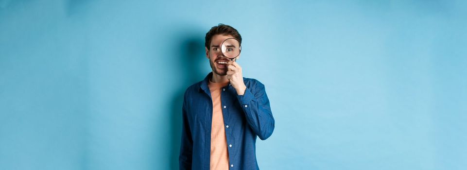 Cheerful young man found something good, smiling and looking through magnifying glass, standing on blue background.