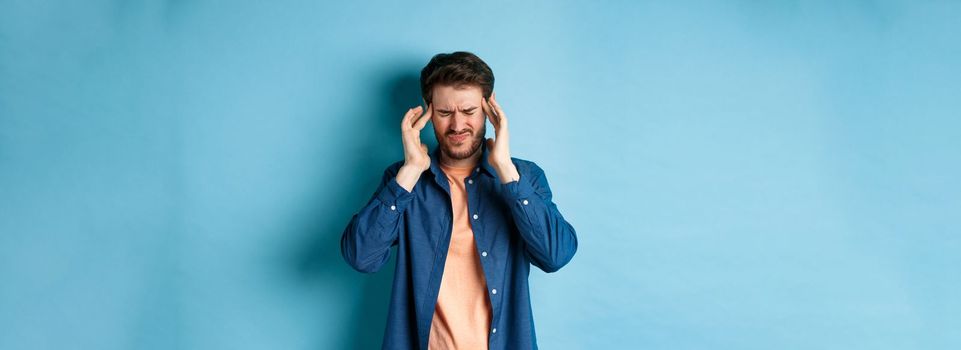 Man feeling sick, touching head and grimacing from painful migraine, having headache or feel dizzy, standing on blue background.