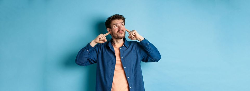 Annoyed young man block sound, shut ears with fingers and grimacing, disturbed by loud neighbours, looking up irritated, standing on blue background.