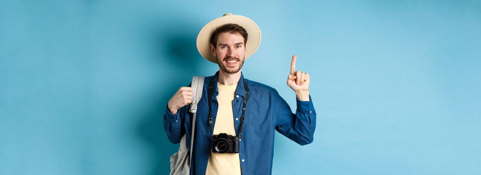 Handsome happy tourist in summer hat, holding backpack and camera, pointing finger up at logo, recommending travel agency or place on vacation, blue background.