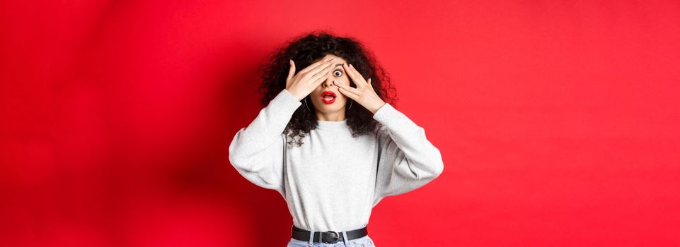 Shocked caucasian girl covering eyes and peek through fingers at something unbelievable, gasping speechless, standing on red background.