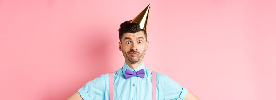 Holidays and celebration concept. Close-up of confused male entertainer in party hat and bow-tie, looking puzzled and shocked, standing over pink background.