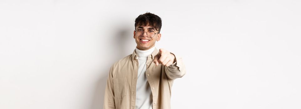 Handsome modern guy in glasses pointing at camera, smiling and choosing you, recruiting or inviting to event, standing on white background.