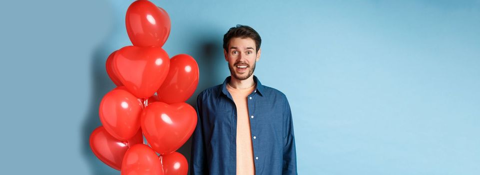 Happy valentines day. Excited smiling guy standing near red hearts balloons and looking at camera, blue background.