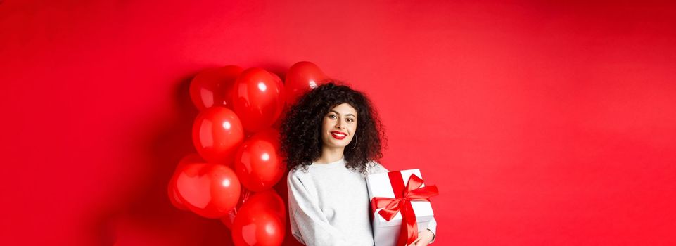 Beautiful caucasian woman with dark curly hair and red lips, standing with romantic hearts balloons and present in box, receive gift from lover, standing on studio background.