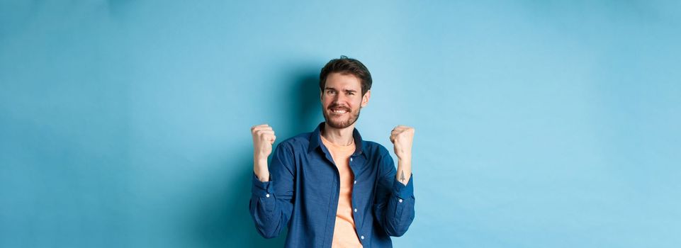Image of happy man celebrating success, shouting yes and shaking clenched fists, achieve goal and triumphing, winning and dancing, standing on blue background.