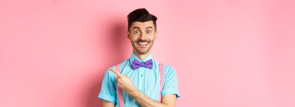 Handsome caucasian guy with bristle and moustache pointing finger right aside, showing advertisement, standing over pink background in classy bow-tie.