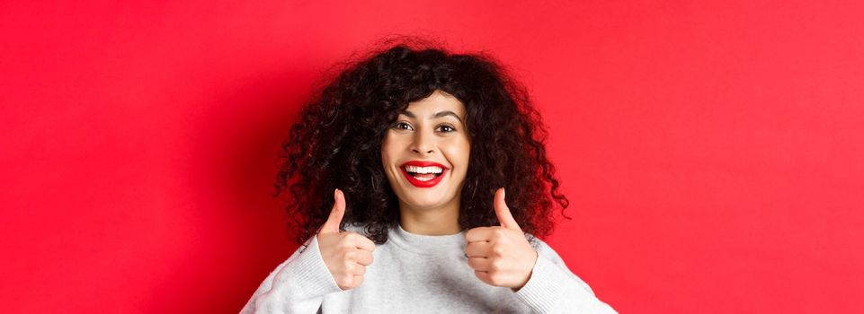 Close up of cheerful caucasian woman showing thumbs up in approval, smiling pleased, like and praise something good, standing on red background.