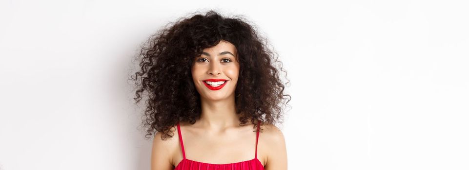 Close-up of beautiful woman with curly hairstyle, wearing red dress and lipstick, smiling happy at camera, white background.