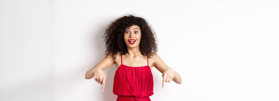 Excited woman with curly hair and red lips, wearing evening dress, gasping and pointing fingers down at super cool promo, standing on white background.