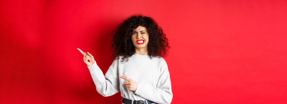 Skeptical caucasian woman grimacing dissasitsfied, complain on bad taste, pointing fingers right and cringe from dislike, standing against red background.