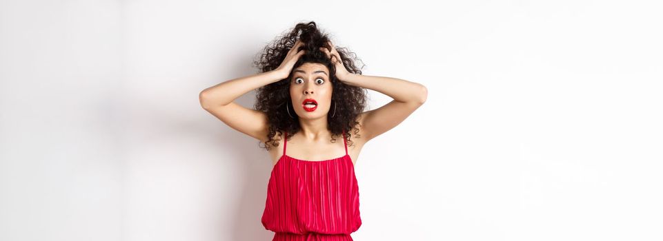 Shocked young woman holding hands on head and panicking, staring frustrated at camera, wearing red dress and makeup, white background.