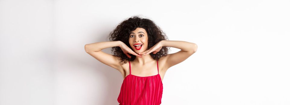 Beauty. Happy elegant woman with curly hair and makeup, wearing red dress, showing her face and smiling excited, standing against studio background.