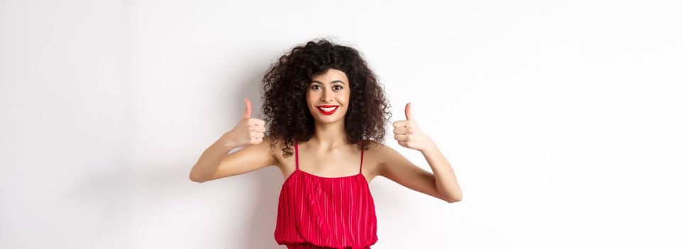 Pretty female model with curly hair and red dress, bright makeup, showing thumbs up in approval, say yes, smiling and nodding in approval, white background.