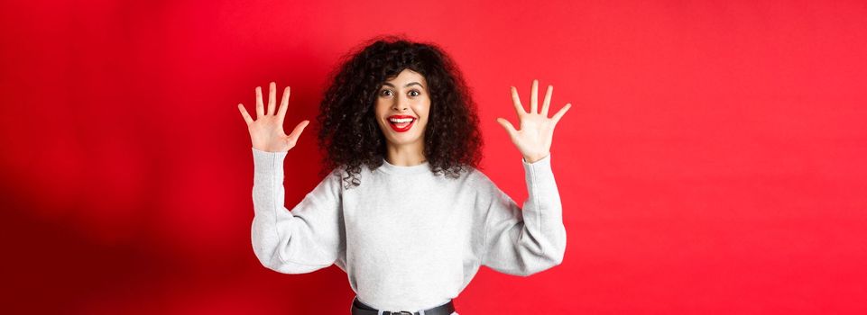 Happy beautiful woman smiling and raising hands up, showing number ten, ordern dozen of something, standing in sweatshirt on red background.