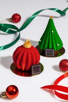 Tempting pastries in shape of Christmas ball and tree covered with red and green sugar glaze on white background with ribbons and baubles. Festive collection of authors desserts