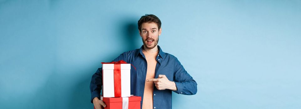 Handsome young man pointing at valentines gift boxes, standing over blue background in casual clothes. Copy space