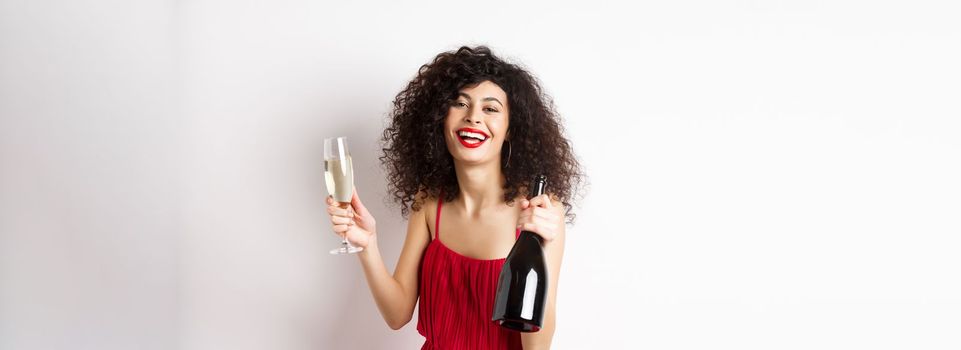 Happy party woman in red dress, laughing and holding bottle of champagne with glass, drinking and having fun, celebrating holiday, standing on white background.