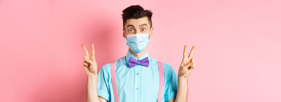 Coronavirus, healthcare and quarantine concept. Funny guy in bow-tie and face mask showing peace, victory signs and looking happy at camera, standing on pink background.