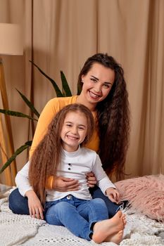 Love of young mother and daughter. They sit on the bed at home and have fun, mother hugs daughter