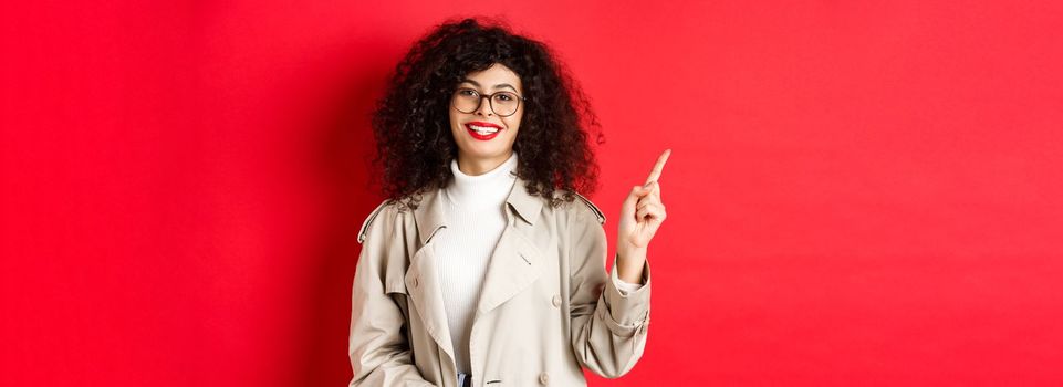 Portrait of stylish caucasian woman in trench coat and glasses pointing finger aside, showing empty space on red background and smiling.