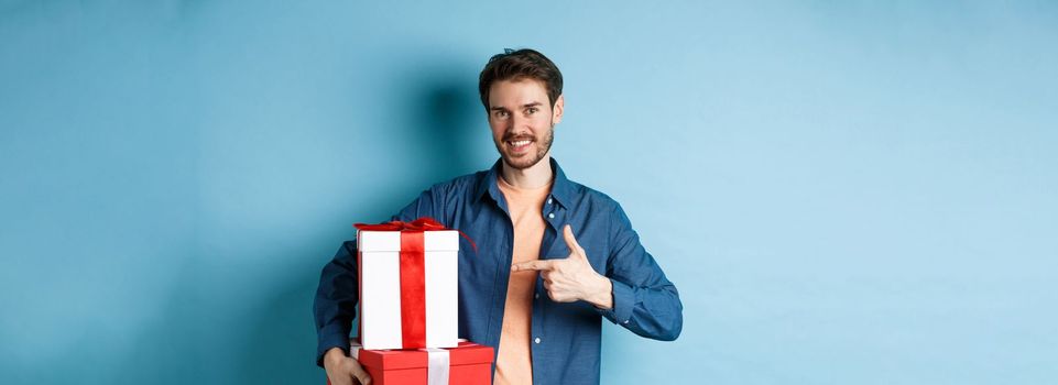 Attractive boyfriend pointing finger at valentines day surprise gifts, showing romantic presents for girlfriend, standing against blue background.
