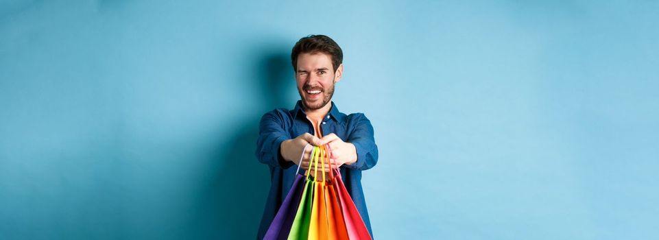Cheerful handsome man winking and smiling, extending hands with colorful shopping bags, standing on blue background.