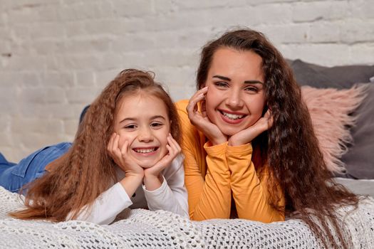 Love of young mother and daughter. They lie on the bed at home and have fun, they lie nearby and look into the camera