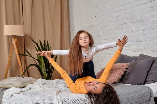 Love of young mother and daughter. They lie on the bed at home and have fun, they are fooling around, mother is holding her daughter on top of her legs